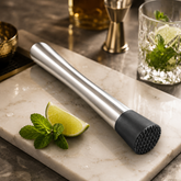 Montavo Cocktail Muddler and Fruit Crusher with stainless steel handle and black textured base beside lime and fresh mint.