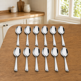 Montavo by FnS Divine 12-piece stainless steel soup spoon set arranged on a wooden table in a bright natural home kitchen.