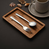 Fork and spoon on wooden tray, styled with seashells and coffee mug on dark background.