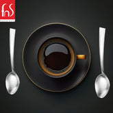 FnS Premium Dining overhead view of black coffee cup with gold rim surrounded by two polished stainless steel teaspoons on dark textured background.
