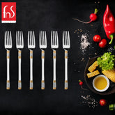 Six stainless steel dinner forks with decorative handles are neatly aligned on a black surface. To the right are fresh vegetables, spices, and a small bowl of sauce. The top left corner has a red logo with white text "fns Premium Dining".