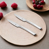 Two sleek Stainlessss steel dinner knives on beige woven placemat with fresh strawberries and red grapes in background setting.