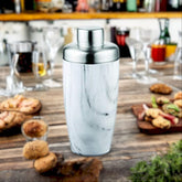 FNS marble finish cocktail shaker on wooden bar table with appetizers, cookies, glassware, and bottles in background