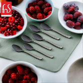 Six black teaspoons with decorative silver geometric handles arranged on green mat, surrounded by white bowls filled with mixed berries and raspberries.