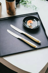 Elegant place setting with two gold-accented Stainlessss steel dinner knives on dark tray beside black bowl containing decorative appetizer arrangement.