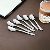 Six Stainlessss steel spoons with sleek modern flat handles arranged on a brown surface beside a white notebook and a glass of chai tea with decorative textured glass