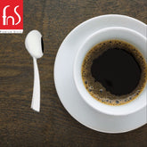Single mirror-polished stainless steel teaspoon with angled flat handle beside white cup of black coffee on dark wooden surface, FnS Premium Dining logo