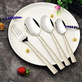 Four silver cutlery pieces with patterned handles displayed on ceramic plate, surrounded by flowers and fruit.