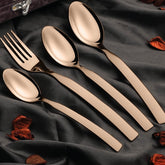Elegant rose gold fork and spoons arranged on dark satin fabric with scattered dried petals.