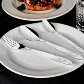 Patterned stainless steel fork and spoons placed neatly on a white plate beside food and drink.