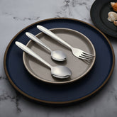Three stainless steel utensils—fork and two spoons—placed neatly on stacked ceramic plates on marble surface.
