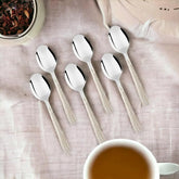 Six stainless steel tea spoons with textured white handles arranged on beige linen fabric with tea cup and dessert bowl