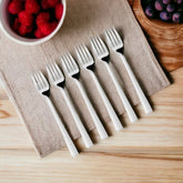 Six white dinner forks with black triangular accents on handles arranged on beige textured placemat with berries nearby