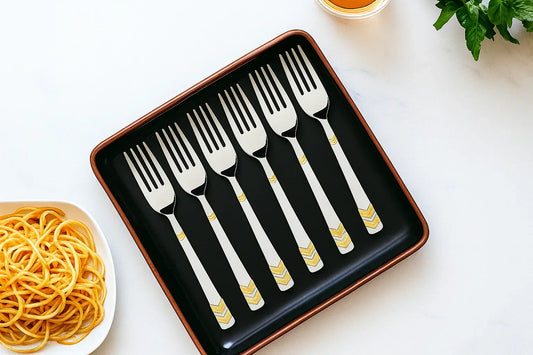 Six silver forks with gold chevron designs on the handles neatly arranged in a black rectangular tray with brown edges, placed on a white surface next to a bowl of spaghetti, a glass of amber liquid, and green herbs.