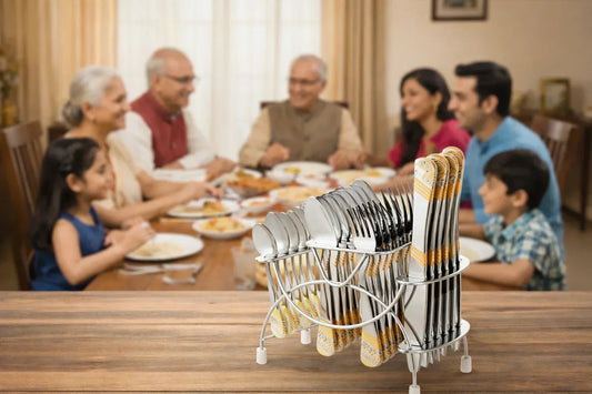 A metal cutlery holder filled with neatly arranged forks, spoons, and knives sits on a wooden table in the foreground, while a multigenerational family smiles and eats dinner together in a softly blurred background dining room.