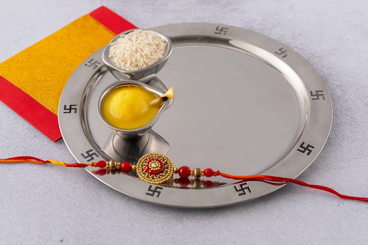 Traditional stainless steel pooja thali with swastik design, containing a lit ghee diya, rice bowl, and decorative rakhi, placed on a festive yellow and red cloth.