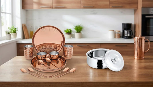 Stylish kitchen setup featuring a copper Indian dinner set with engraved thali plate, bowls, glasses, spoons on wooden table, beside a shiny stainless steel casserole with lid, modern wooden cabinets background.
