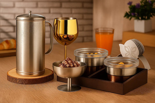 Modern stainless steel serveware set with insulated jug, golden wine glass, bowls of snacks, and nut holder on a wooden dining table.