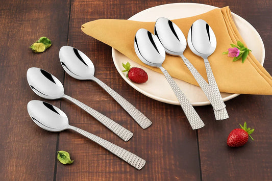 A set of six stainless steel spoons with textured handles placed on a wooden surface beside a plate with a folded napkin and strawberries.