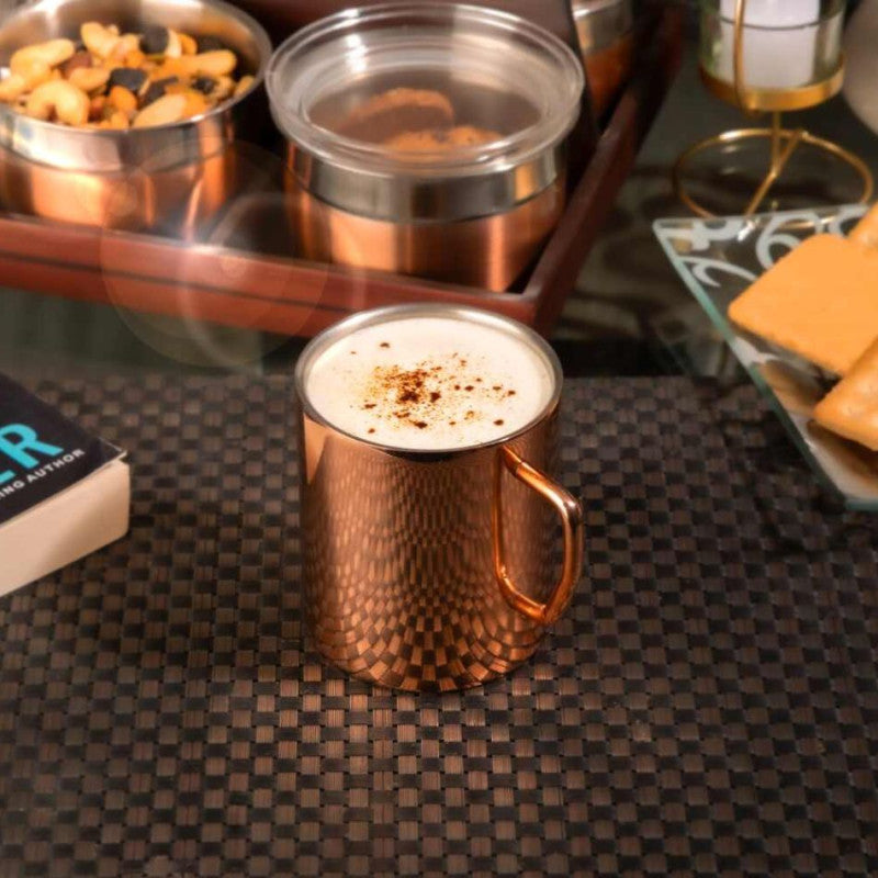 Close-up of copper hammered mug filled with frothy cappuccino on checkered placemat with serving tray in background