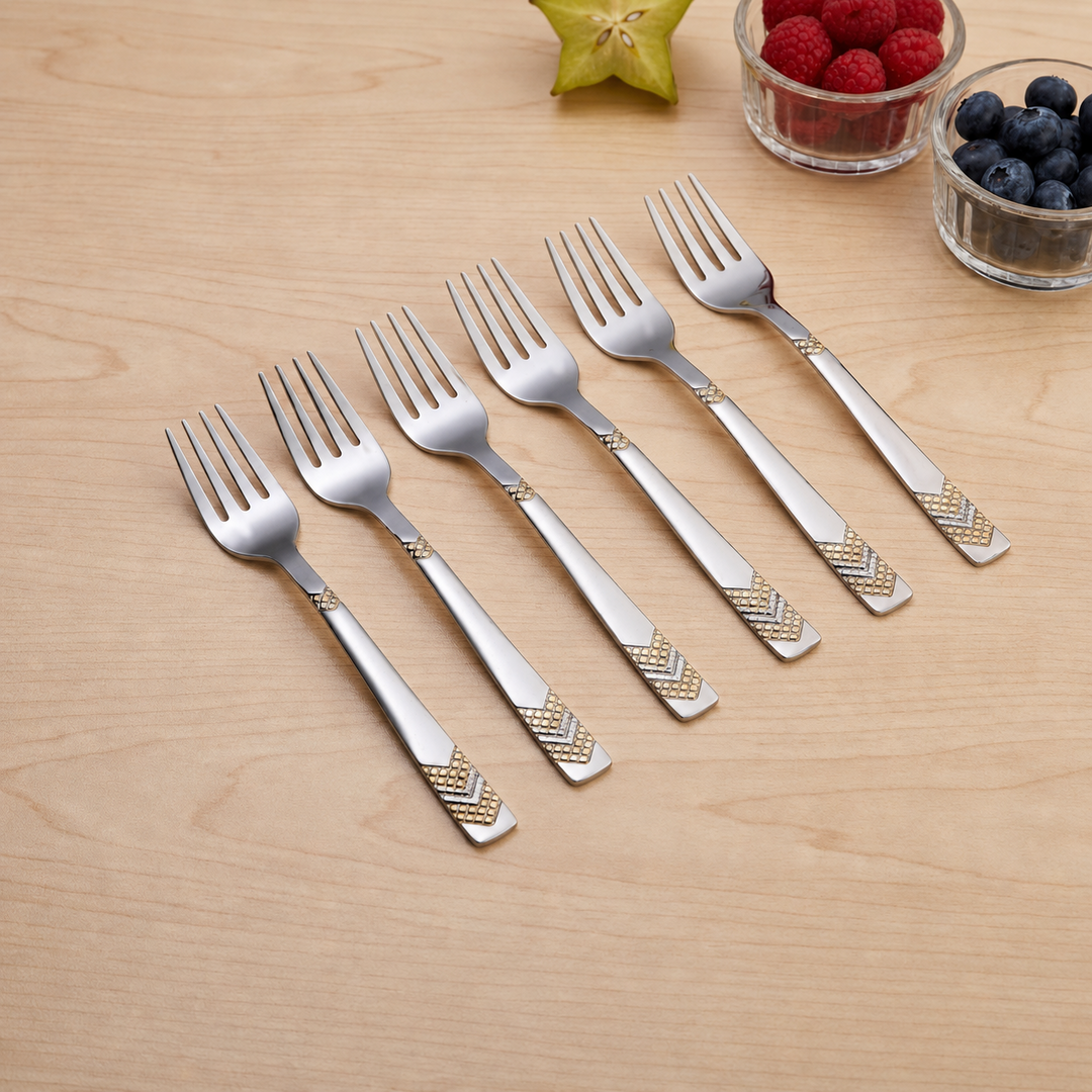 Six stainless steel fruit forks scattered on beige surface near pastries, displaying chevron gold pattern design details