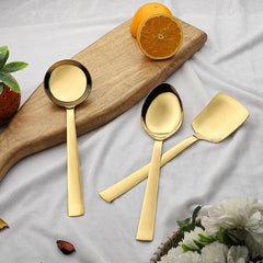 Three gold serving spoons including gravy ladle and rice server displayed on wooden board with fresh orange slices
