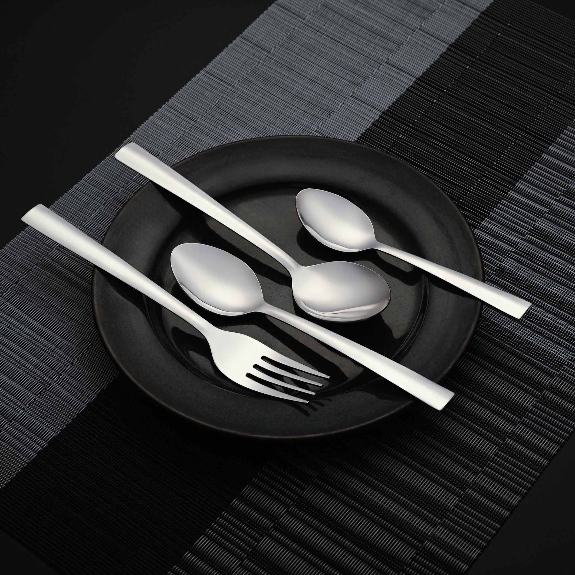 Minimalist stainless steel cutlery neatly placed on a black ceramic plate over a woven black mat.