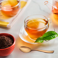 Glass teacup with tea and fresh leaves beside a single rose gold spoon on marble surface.