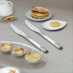 Two stainless steel dinner knives with decorative gold and silver bands on handles, displayed on breakfast table with pancakes and eggs.