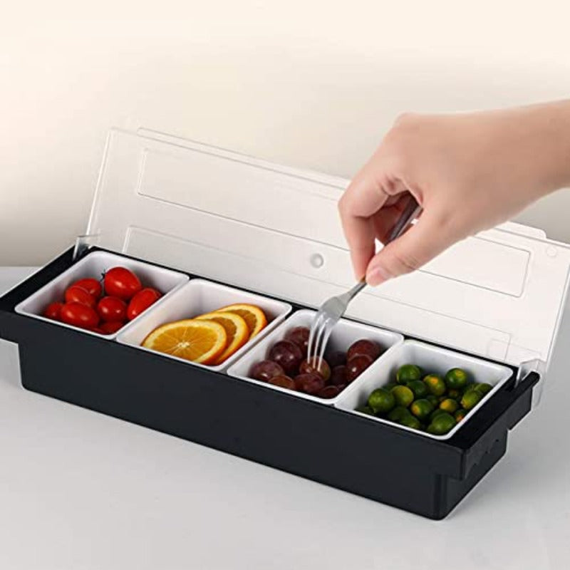 Black condiment caddy with four white compartments filled with cherry tomatoes, orange slices, grapes, and olives with clear hinged lid and hand using tongs