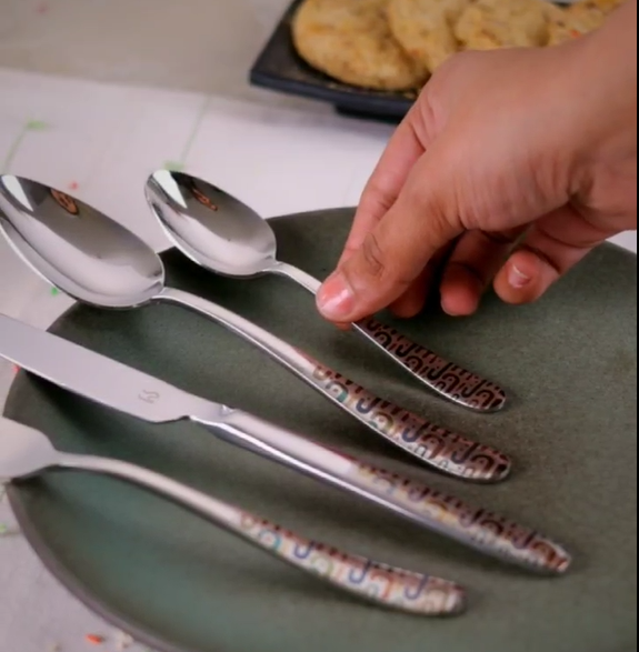 Hand reaching for a spoon from a cutlery set with decorative handles, placed on a plate.