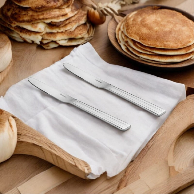 Two stainless steel Flair dinner knives with triple-line handle detail on white napkin beside wooden board with stacked pancakes.