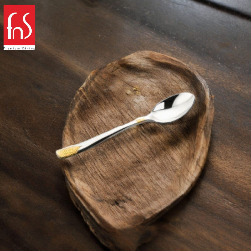 Single stainless steel teaspoon with gold-textured handle tip placed diagonally on rustic wooden heart-shaped dish, FnS Premium Dining logo in corner