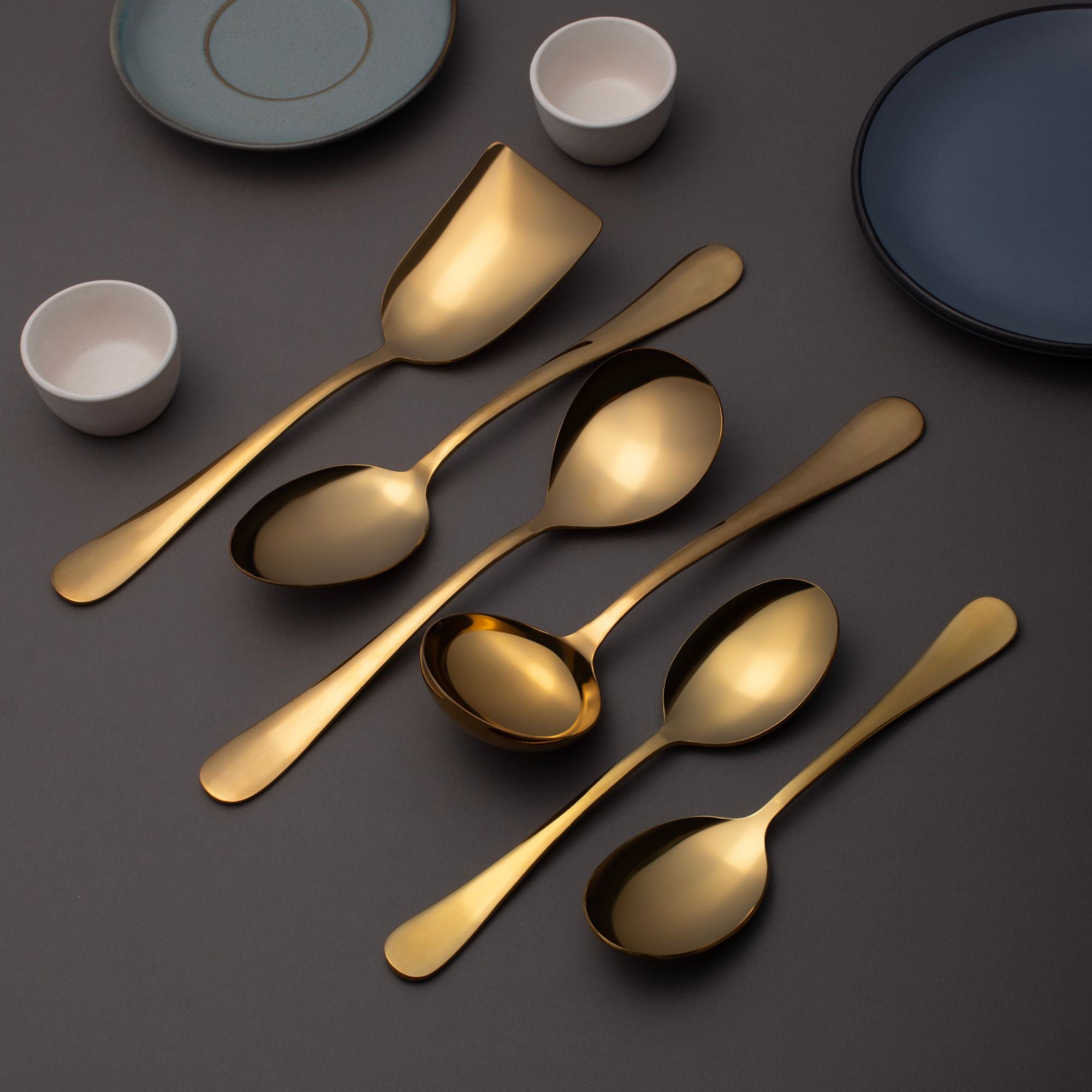 FNS Aurik gold plated stainless steel 6-piece serving set arranged diagonally on dark gray surface with modern ceramic dinnerware and white bowls