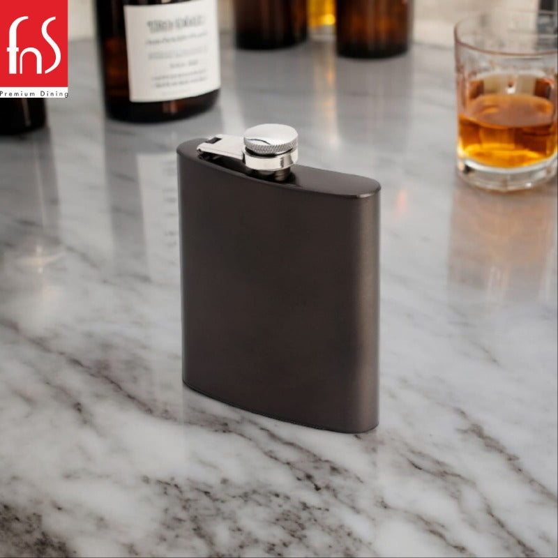 FNS Premium Dining black finish hip flask with stainless steel screw cap on white marble counter with liquor bottles and whiskey glass in background