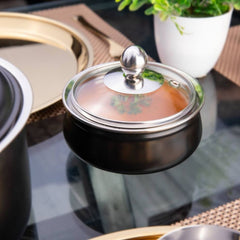 FNS black stainless steel handi with closed glass lid, side-angle view on a reflective table with gold dinnerware and plant in background