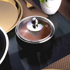 FNS black stainless steel handi with closed transparent glass lid, top-angle close-up view on a reflective dining table surface