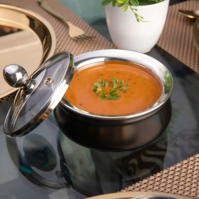FNS black stainless steel handi with glass lid placed aside, filled with tomato soup topped with fresh coriander garnish, styled on an elegant dining table setting