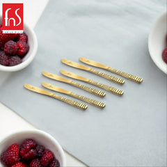 Styled flat lay of six gold Boston dinner knives with textured handles arranged on light blue surface, surrounded by white bowls filled with fresh red raspberries and premium dining brand logo.