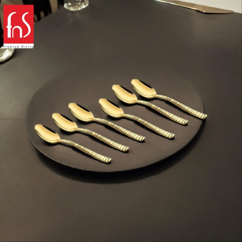 Six gold-plated teaspoons with textured handles arranged on a dark round plate in an upscale dining setting with glassware visible.