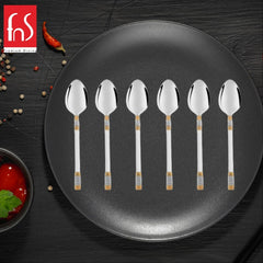 FNS Celebration 6-piece baby spoon set with 24-karat gold-silver pattern handles displayed on dark plate with red chili and spices, premium infant feeding cutlery.