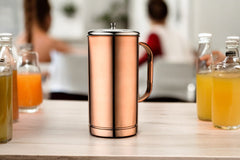 Rose gold lidded stainless steel water jug with three two-tone copper-silver tumblers arranged on a rustic stone surface with a natural outdoor green background.