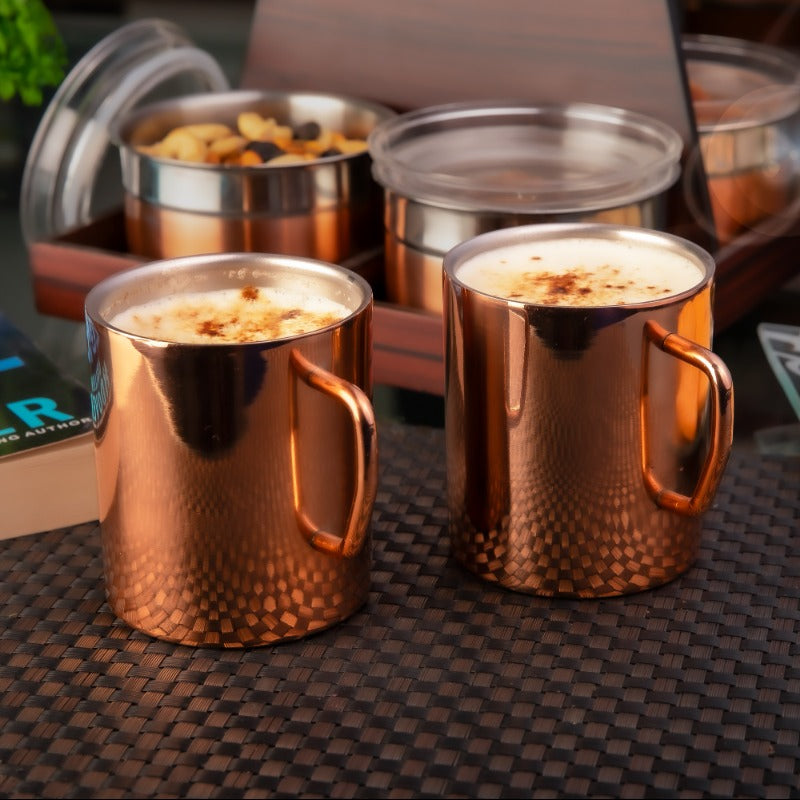 Two copper-finished stainless steel coffee mugs with hammered texture pattern, filled with frothy cappuccino topped with cinnamon powder, placed on a dark woven placemat with snack containers in the background.
