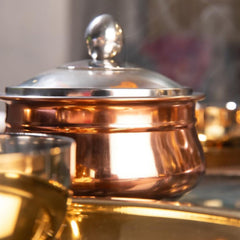Close-up detail of fns copper finish stainless steel handi with glass lid and steel knob, styled alongside gold cookware in warm ambient light