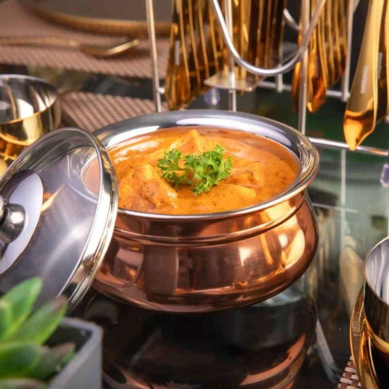 fns copper finish stainless steel handi with lid open, filled with butter chicken curry garnished with parsley, styled on dark surface with gold cutlery in the background