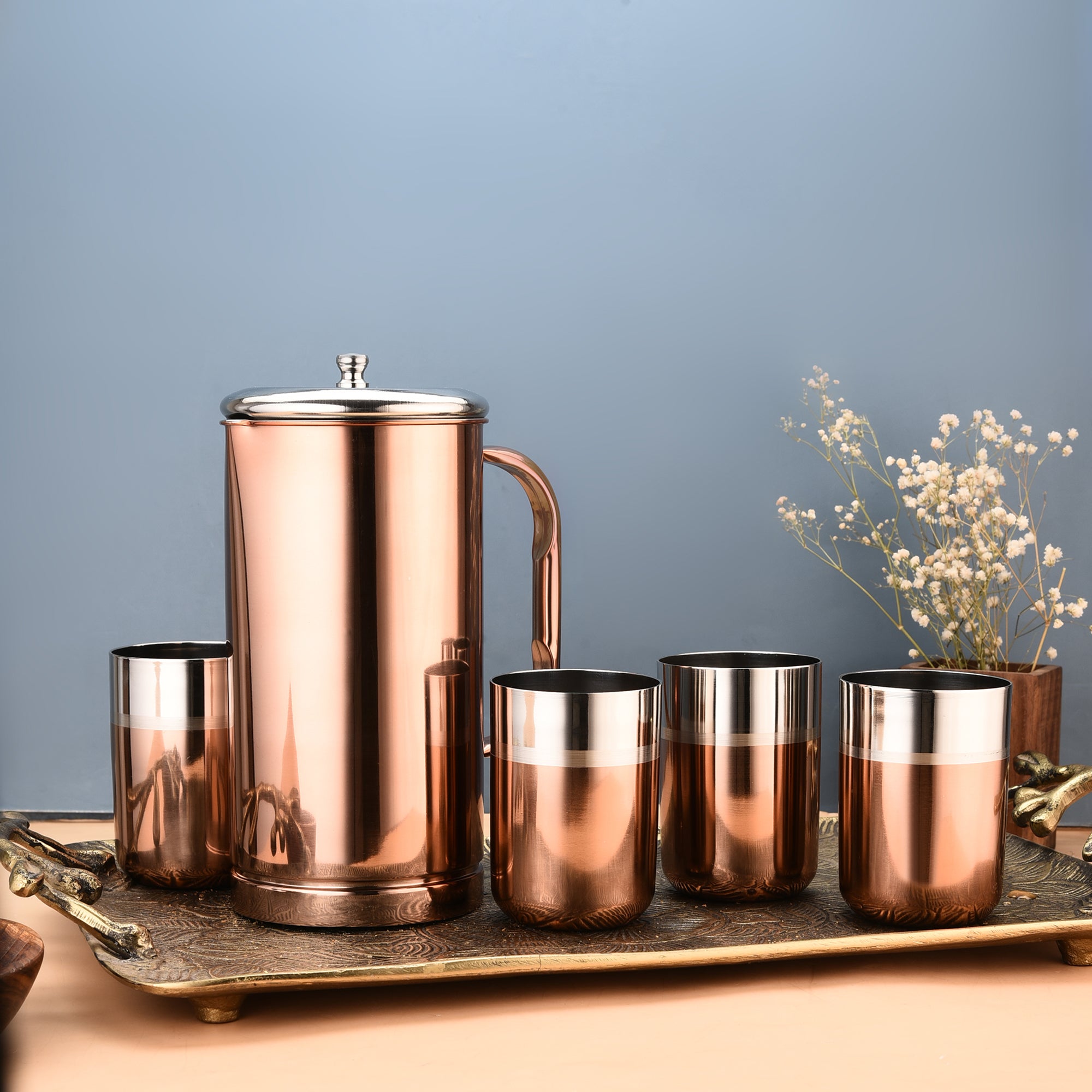 Rose gold stainless steel water jug set with lidded pitcher and four two-tone copper-silver tumblers, styled on a decorative tray with dried white flowers against a blue background.
