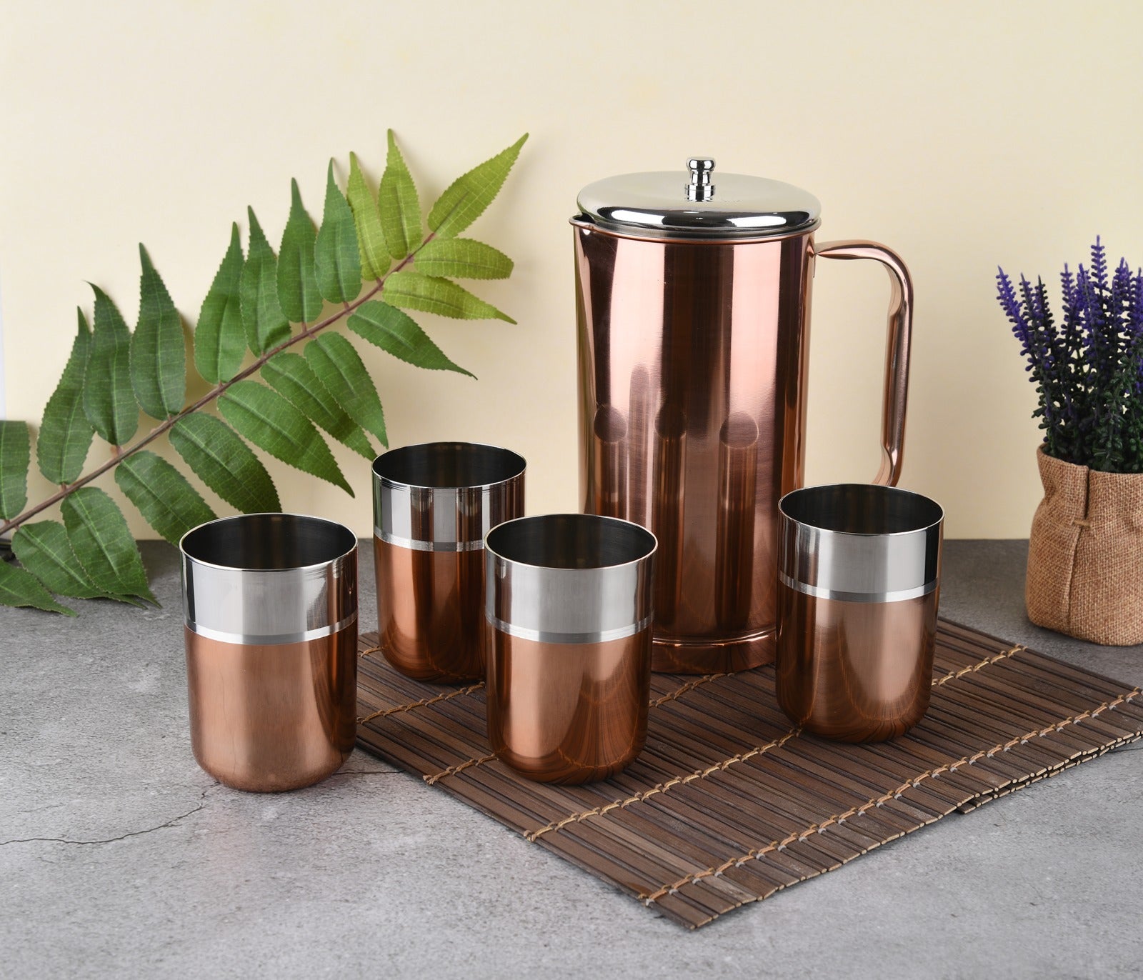 Rose gold stainless steel water jug with silver lid and handle, alongside three two-tone copper-silver cylindrical tumblers including one tipped on its side, on white background.