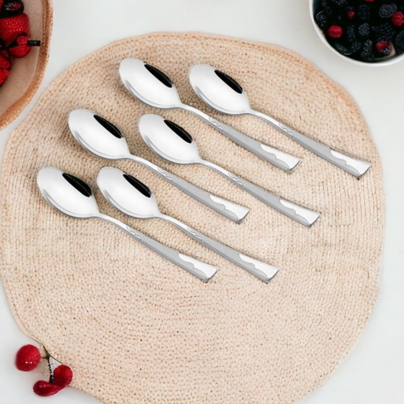 Six polished stainless steel teaspoons arranged on a natural woven placemat, surrounded by fresh berries in bowls and scattered raspberries and blackberries.