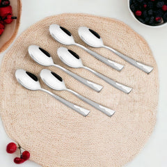 Six polished stainless steel teaspoons arranged on a natural woven placemat, surrounded by fresh berries in bowls and scattered raspberries and blackberries.