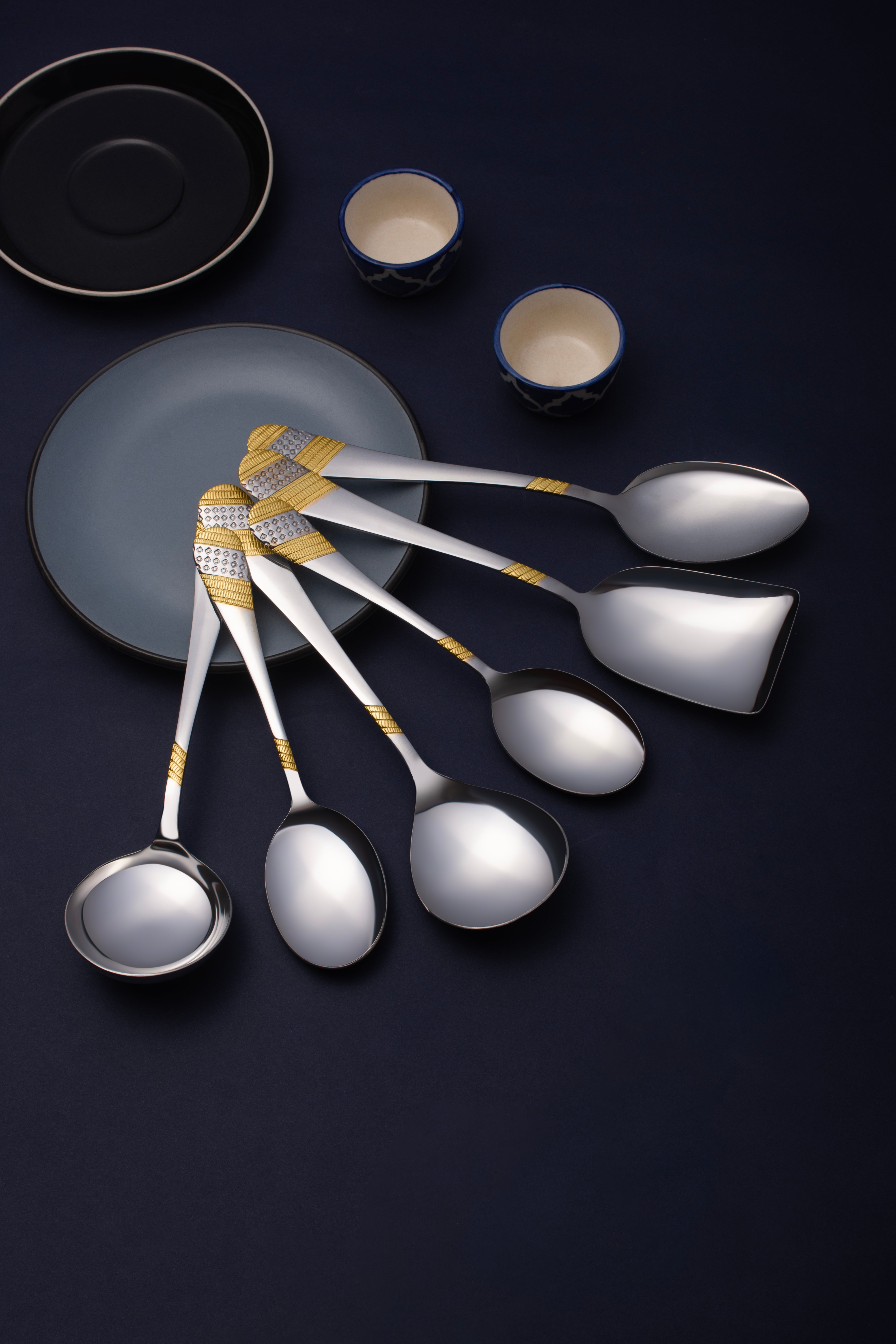 Seven-piece serving spoon set with gold-embellished handles fanned over blue-black table setting with plates.